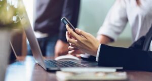 man's hand using a smartphone with a laptop in front of him